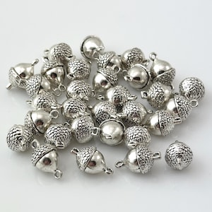 May include: A collection of silver-toned metal acorn charms, each featuring a textured cap with a detailed scale pattern and a smooth, rounded base. Every charm includes a small metal loop at the top for attachment, scattered across a plain surface.
