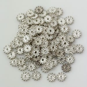 May include: A pile of silver-toned metal bead caps with a decorative, scalloped edge. The bead caps are small and round, with a hole in the center for threading.