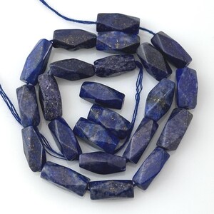 May include: A strand of faceted, elongated hexagonal beads made of deep blue lapis lazuli stone. Each bead features natural gold-colored pyrite inclusions and is strung on a thin blue thread, arranged in a loose, circular coil pattern.