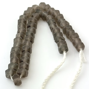 May include: Strands of dark gray, faceted beads strung on white cord. The beads have a hexagonal shape and a slightly frosted appearance. The beads are likely for jewelry making or crafting.