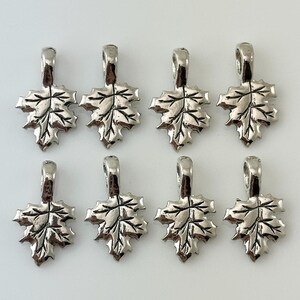 May include: Eight silver-colored maple leaf charms with detailed veining and a loop for attaching to jewelry. The charms are arranged in two rows of four, perfect for crafting necklaces, bracelets, or earrings.