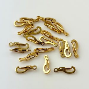May include: A collection of small, gold-colored spring snap hooks, scattered on a white surface. The hooks have a curved design with a spring-loaded clasp. These are likely used for attaching items.