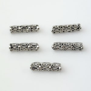 May include: Five silver-colored, rectangular beads with an ornate, openwork design. Each bead features a repeating pattern of curved lines and small circular details, creating a textured appearance. The beads are scattered on a white surface.