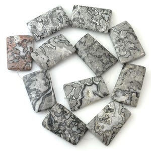 May include: A collection of rectangular gemstone beads in shades of gray, white, and a touch of reddish-brown. The beads have a marbled appearance with intricate veining patterns. These beads are strung together, ready for jewelry making.