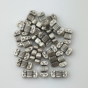 May include: A pile of silver metal beads with a decorative swirl design. The beads are about 1 cm long and have a slightly textured surface.