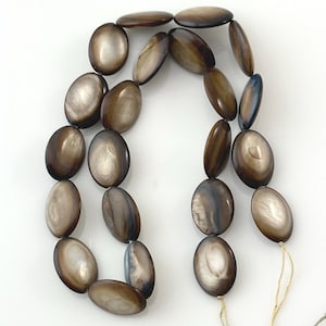 May include: A strand of oval-shaped beads with a smooth, iridescent finish in shades of brown, tan, and cream. The beads are arranged in a loose, curved formation, showcasing a reflective, pearlescent surface texture throughout the entire collection.