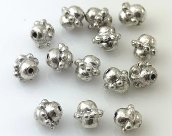 May include: A collection of silver-colored metal beads with an irregular, textured surface. Each bead has a unique, organic shape with small, protruding details and a central hole for stringing. The beads are scattered on a white surface.