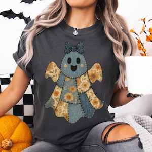 以下が含まれることがあります： パッチワークのゴーストデザインが特徴のダークグレーのTシャツ。ゴーストは青い体、花柄の羽、青いリボンをしています。デザインはシャツに縫い付けられており、ユニークで手作りの外観を作り出しています。シャツは快適でカジュアルなスタイルです。