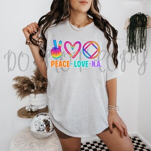 Puede incluir: Una camiseta gris con un signo de paz, un corazón y un círculo con un diamante en su interior. El gráfico está en colores tie-dye arcoíris. El texto "Peace Love N.A." está debajo del gráfico.
