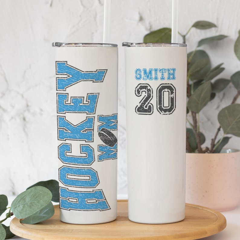 May include: Two white stainless steel tumblers with lids and straws. One tumbler has the text "Hockey Mom" in blue with a hockey puck graphic. The other tumbler has the text "Smith 20" in black.