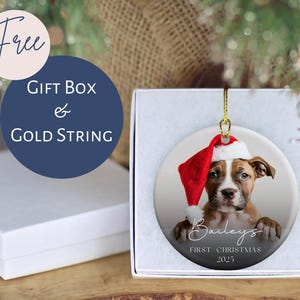 May include: A round Christmas ornament featuring a puppy wearing a Santa hat. The ornament is in a white gift box with a gold string. Text on the ornament reads "Bailey's First Christmas 2025". A blue circle with the words "Gift Box & Gold String" is also visible.