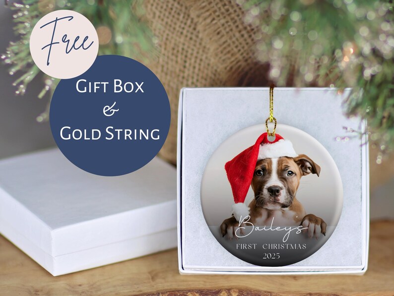 May include: A round Christmas ornament featuring a puppy wearing a Santa hat. The ornament is in a white gift box with a gold string. Text on the ornament reads "Bailey's First Christmas 2025". A blue circle with the words "Gift Box & Gold String" is also visible.