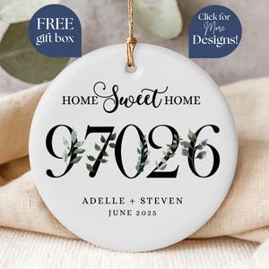 May include: A white ceramic ornament with the words "Home Sweet Home" and the numbers "97026" in black. The numbers are decorated with green and gray leaves. The names "Adelle + Steven" and the date "June 2025" are below. Includes the text "Free gift box" and "Click for More Designs!"