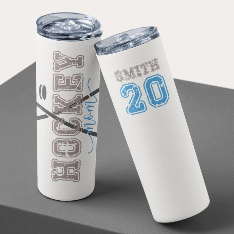 May include: Two white stainless steel tumblers with a hockey theme. One tumbler has the word "HOCKEY" in a distressed font with a hockey stick and puck graphic. The other tumbler has the text "SMITH 20" in a blue distressed font.