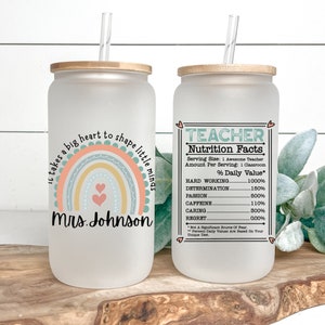 Puede incluir: Un vaso de vidrio esmerilado con tapa de bamb&uacute; y una pajita. El vaso tiene un dise&ntilde;o de arco&iacute;ris con el texto "Se necesita un gran coraz&oacute;n para moldear las mentes peque&ntilde;as" y "Sra. Johnson". El vaso est&aacute; al lado de un vaso similar con el texto "TEACHER Nutrition Facts" y una lista de porcentajes para "Trabajo duro", "Determinaci&oacute;n", "Pasi&oacute;n", "Cafe&iacute;na", "Cuidado" y "Arrepentimiento".