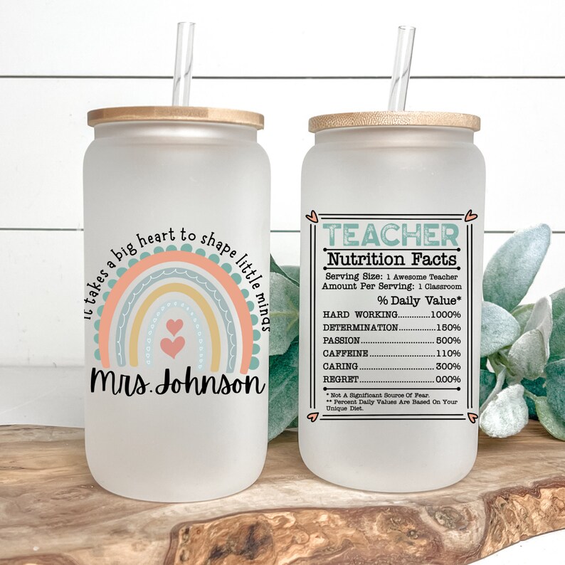Puede incluir: Un vaso de vidrio esmerilado con tapa de bamb&uacute; y una pajita. El vaso tiene un dise&ntilde;o de arco&iacute;ris con el texto "Se necesita un gran coraz&oacute;n para moldear las mentes peque&ntilde;as" y "Sra. Johnson". El vaso est&aacute; al lado de un vaso similar con el texto "TEACHER Nutrition Facts" y una lista de porcentajes para "Trabajo duro", "Determinaci&oacute;n", "Pasi&oacute;n", "Cafe&iacute;na", "Cuidado" y "Arrepentimiento".