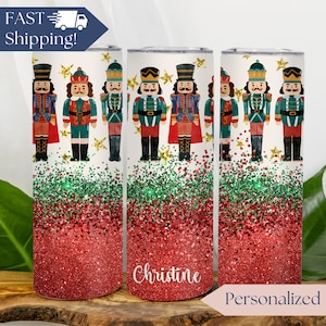 May include: Three stainless steel tumblers with a red and green glitter design. The tumblers feature a Christmas nutcracker design with gold stars. One tumbler has the name "Christine" on it. The tumblers are personalized.