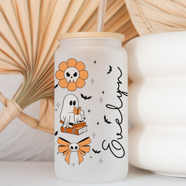 Op de afbeelding: Een frosted glazen beker met een bamboe deksel en rietje. De beker heeft oranje en zwarte Halloween-thema graphics, waaronder een schedelbloem, een spook dat een boek leest en een strik met een schedel. De naam "Evelyn" is in een script geschreven.