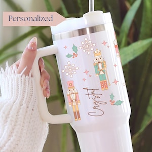 May include: White insulated tumbler with a handle, decorated with nutcracker illustrations, snowflakes, and holly. The word "Personalized" is in a pink banner. The name "Crystal" is written in cursive. A festive holiday theme.