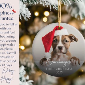 May include: A round Christmas ornament featuring a puppy wearing a Santa hat. The ornament has the text "Bailey's First Christmas 2025". The background is a blurred image of a Christmas tree.