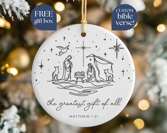 May include: White ceramic Christmas ornament with a black line drawing of the Nativity scene. The text "the greatest gift of all" and "Matthew 1:21" are below the image. Includes a gold string for hanging.