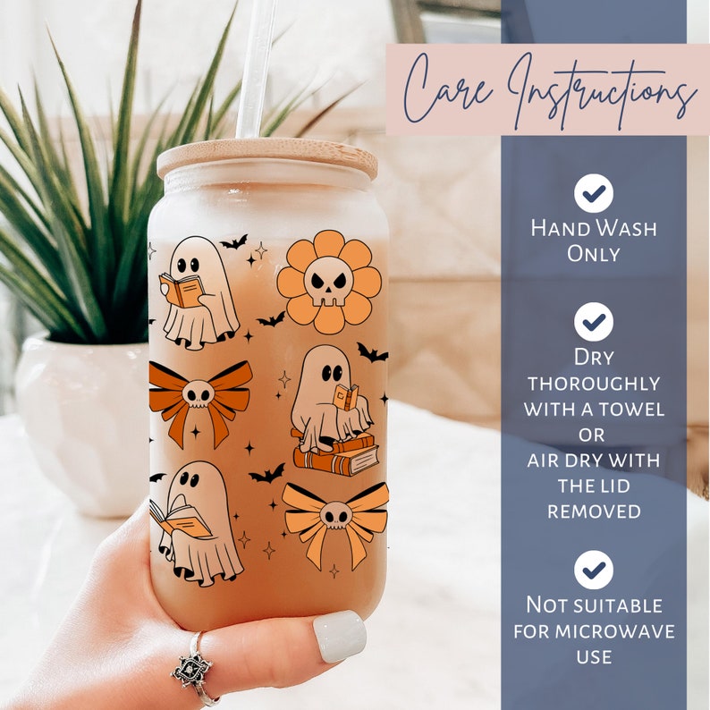 Op de afbeelding: Een glazen beker met een bamboe deksel en rietje, versierd met Halloween-thema illustraties. Het ontwerp omvat geesten die boeken lezen, oranje strikken met schedels, een bloem met een schedel in het midden en vleermuizen. De beker heeft de tekst "Care Instructions".
