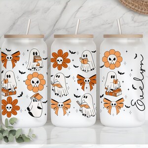 Op de afbeelding: Drie glazen bekers met houten deksels en rietjes. Elke beker heeft een Halloween-thema met geesten, oranje bloemen met schedelcentra en strikken. Het ontwerp bevat vleermuizen en sterren. E&eacute;n beker heeft de naam "Evelyn" in cursief.