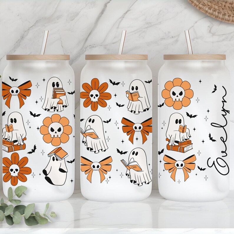 Op de afbeelding: Drie glazen bekers met houten deksels en rietjes. Elke beker heeft een Halloween-thema met geesten, oranje bloemen met schedelcentra en strikken. Het ontwerp bevat vleermuizen en sterren. E&eacute;n beker heeft de naam "Evelyn" in cursief.