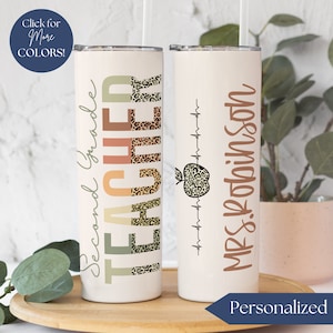 May include: Two white stainless steel tumblers with a personalized design. The first tumbler has the text "Second Grade Teacher" with a leopard print design. The second tumbler has the text "Mrs. Robinson" with a red apple and a heartbeat graphic.