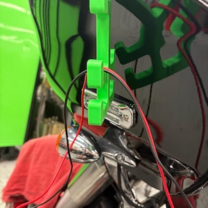 May include: A close-up of a motorcycle with a black and green helmet. A bright green 3D-printed bracket holds a small light and red and black wires. The motorcycle's chrome handlebars and headlight are visible.