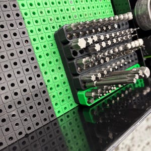 May include: A close-up shot of a tool organizer with various bits. The organizer features black and green plastic components with multiple holes. The bits are silver and arranged in rows. The overall theme is tools and organization.