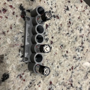 May include: A gray plastic battery holder with the text "TECH ANGLE LIFE SUPPORT" printed on the side. The holder contains several AA batteries. The holder is designed to be mounted on a wall or other surface.