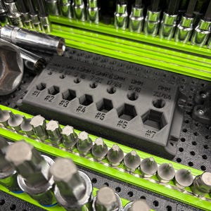 May include: A dark gray 3D-printed plastic organizer block featuring seven numbered slots and various size labels including 5/32, 3/16, 7/32, 17/64, 5/16, and 23/64. The organizer is positioned among rows of silver metal tool bits and sockets on a green tray.