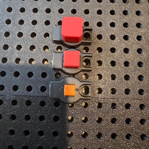 May include: Three small, dark gray plastic components arranged vertically. Each piece features a square button on top, with the top two buttons being red and the bottom button being orange. Each component has a circular hole on one side.