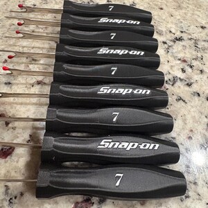 May include: A set of black Snap-on tools with silver shafts and red tips. Each tool has the number "7" and the "Snap-on" logo in white. The tools are arranged in a neat, organized row.