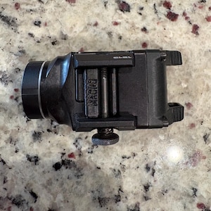 May include: A black tactical light with a cylindrical front and a rectangular body. The light has a mounting bracket and a textured adjustment knob. The word "MACRO" is visible on the device.