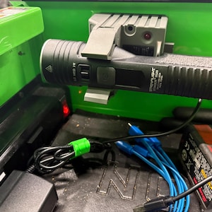 May include: A black and gray Streamlight Stinger 2020 flashlight is mounted in a green toolbox. The flashlight is next to a charging base. A black power adapter, blue cables, and a black cord with a green tie are also visible.