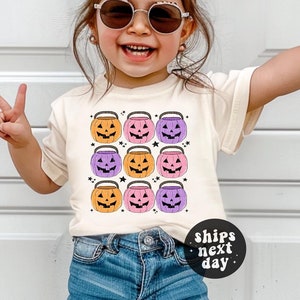 May include: A young child wearing a white t-shirt with a pattern of orange, pink, and purple jack-o'-lantern buckets. The shirt has a small black circle with the text "ships next day". The child is wearing blue denim shorts and sunglasses.