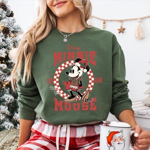 A woman wearing a green sweatshirt with a Minnie Mouse graphic and holding a mug with a Santa Claus design.