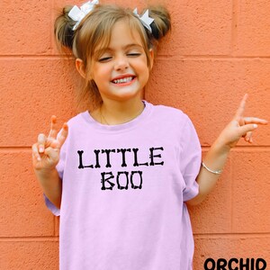 A young girl with pigtails is smiling and making a peace sign with her hands, wearing a pink t-shirt with the text "LITTLE BOO" printed on it.