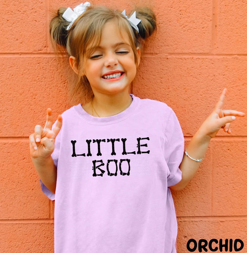 A young girl with pigtails is smiling and making a peace sign with her hands, wearing a pink t-shirt with the text "LITTLE BOO" printed on it.