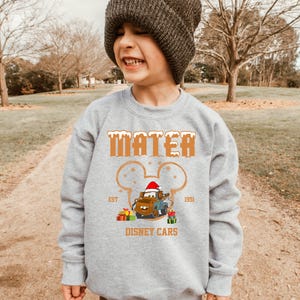 A young boy wearing a gray sweatshirt with a cartoon character and text printed on it, standing in a park.
