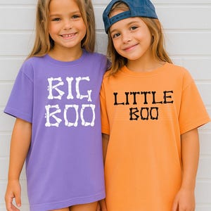 Two young girls wearing purple and orange t-shirts with the text "BIG BOO" and "LITTLE BOO" printed on them, respectively.