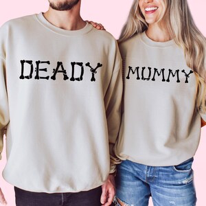 A man and a woman standing side by side, both wearing matching beige sweatshirts with the word "DEADY" printed on them.