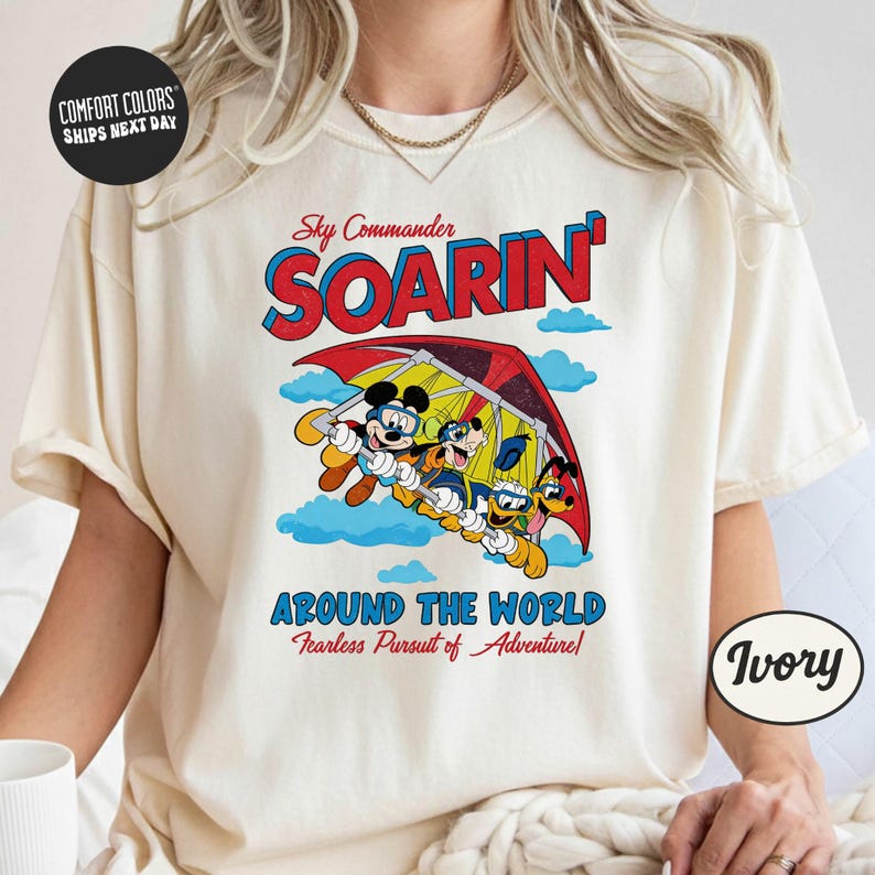A white t-shirt with a colorful graphic design depicting Mickey Mouse and Donald Duck on a hot air balloon, along with the text "Sky Commander SOARIN' Around the World Fearless Pursuit of Adventures".