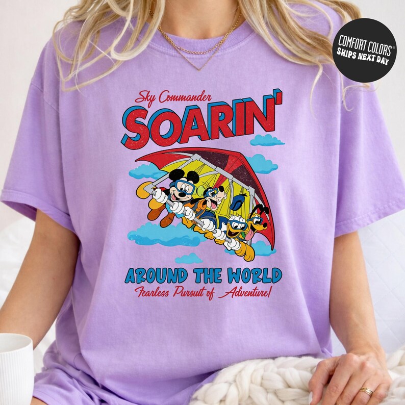 A person wearing a purple t-shirt with a graphic design featuring a cartoon character and the text "Soarin' Around the World Fearless Pursuit of Adventure!"