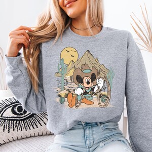 a woman sitting on a bed wearing a mickey mouse sweatshirt