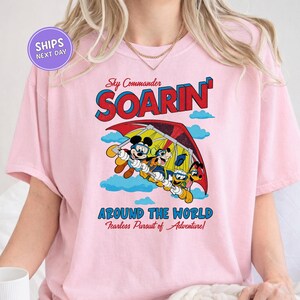 A pink t-shirt with a graphic design featuring a cartoon character and the text "Soarin' Around the World Fearless Pursuit of Adventure!"