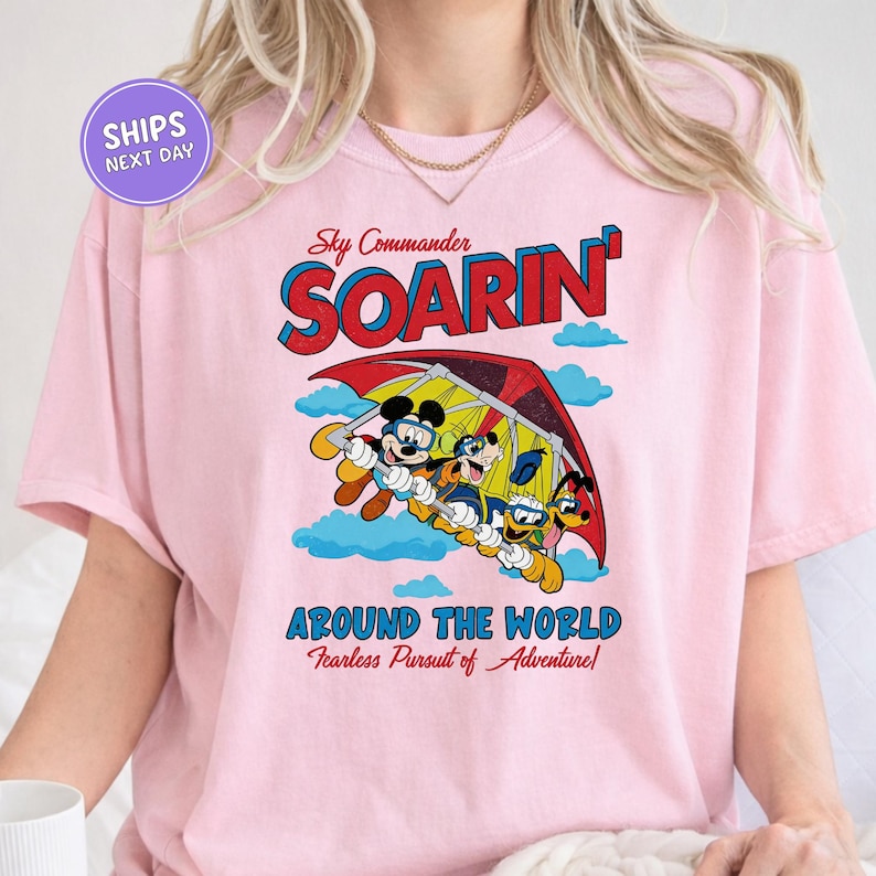 A pink t-shirt with a graphic design featuring a cartoon character and the text "Soarin' Around the World Fearless Pursuit of Adventure!"