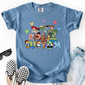 A blue t-shirt with a colorful graphic of characters from the Toy Story franchise, including Woody, Buzz Lightyear, and other beloved characters, along with the word "CUSTOM" written on it.